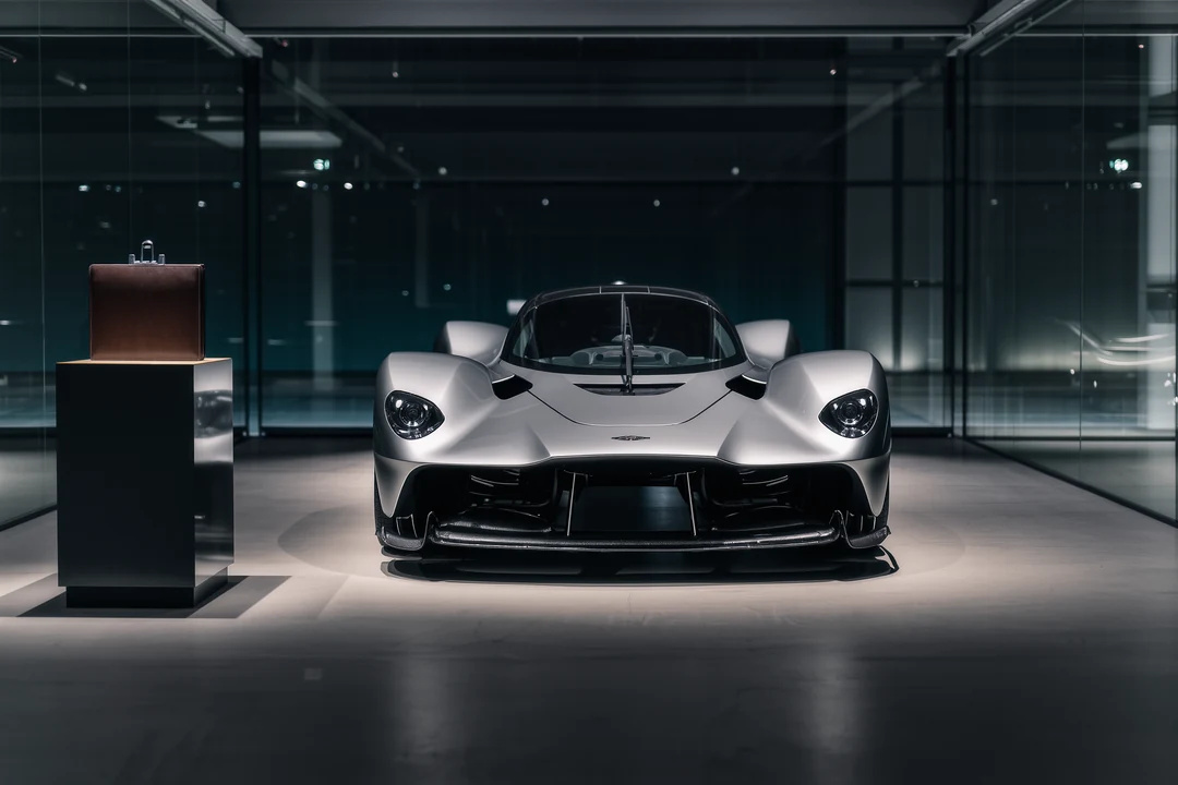 A stripped, damaged car missing its wheels, doors, and rear hatch sits abandoned on rough ground near other vehicles—far from the sleek precision of hypercars.