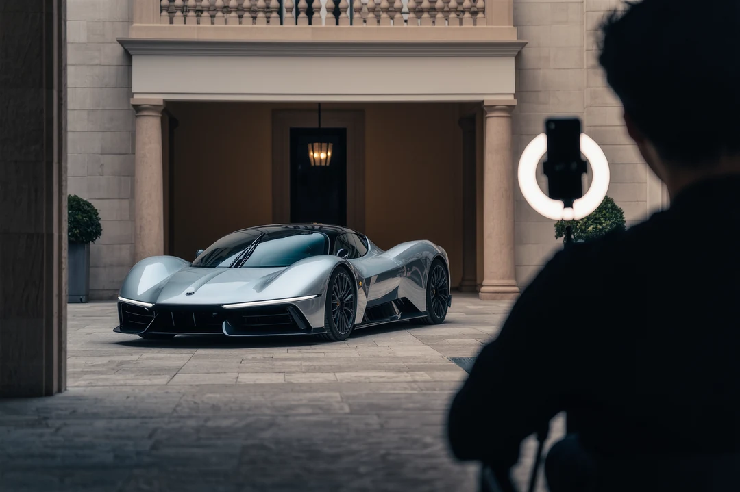 A man in a suit stands by an open hypercar door, pointing upward, in front of a building with a sign reading "HORRAK.