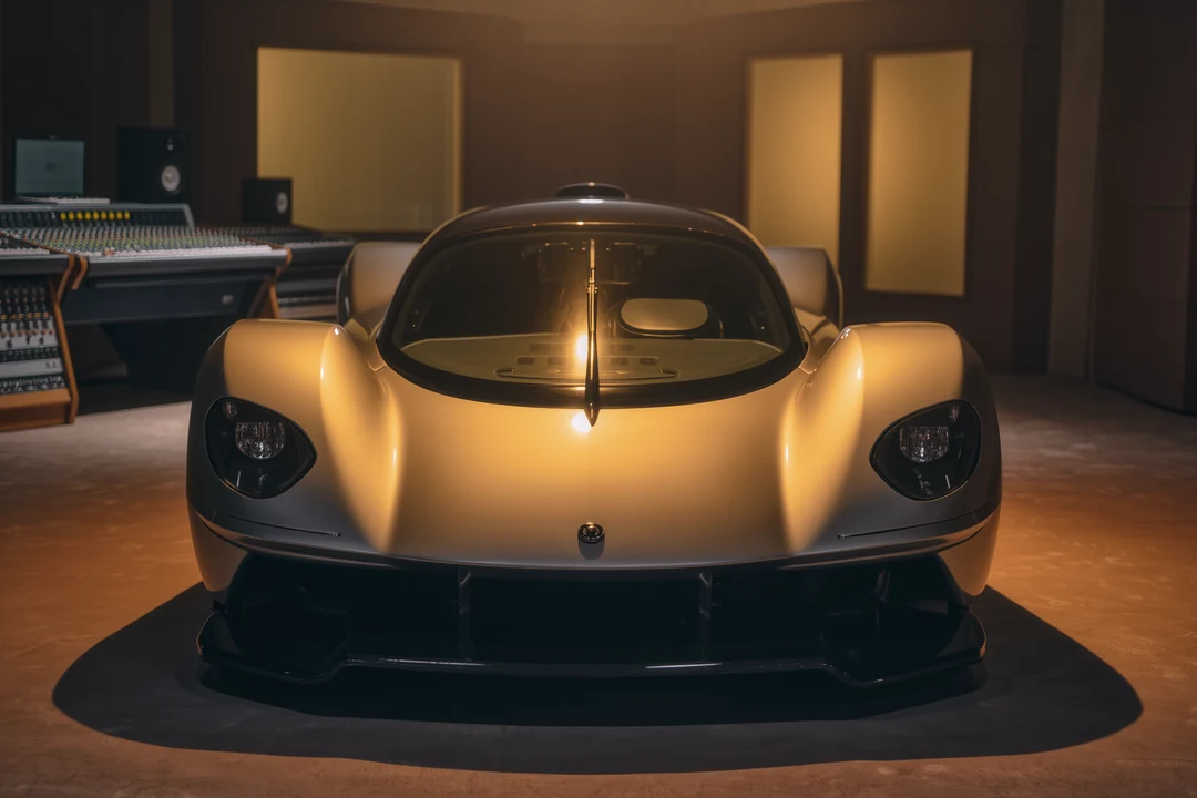 Close-up of the front left side of a black Porsche hypercar, showing a headlight, part of the hood with raindrops, and the Porsche logo, parked on cobblestone pavement.